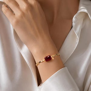 May include: A gold-toned bracelet featuring a single rectangular deep red gemstone at the center. The band is composed of smooth, polished metal links that connect to the central stone setting, creating a refined and minimalist jewelry piece.