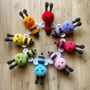 Pode incluir: Oito brinquedos de abelha em crochê dispostos em círculo. Cada abelha apresenta uma cabeça redonda, um corpo listrado em várias cores incluindo vermelho, laranja, amarelo, verde, azul, roxo, rosa e rosa claro, asas brancas e duas antenas.