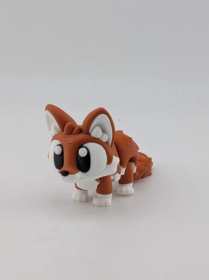 Peut inclure: Une petite figurine de renard articul&eacute;e pr&eacute;sentant un corps orange vif avec des accents blancs sur la poitrine, les pattes et l'int&eacute;rieur des oreilles. Le renard a de grands yeux noirs ronds et un petit nez noir, avec des lignes d'impression 3D visibles.