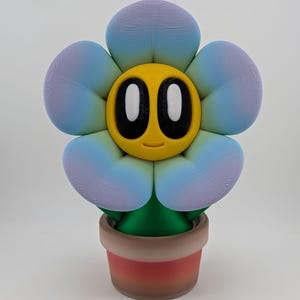 May include: A 3D printed flower figurine featuring five rounded petals with a gradient of blue and purple hues. The center is a yellow circular face with large black oval eyes and a small smile. The flower sits in a small, brown, cylindrical pot.