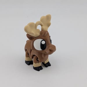 May include: A small, brown, 3D-printed reindeer figurine with large white eyes, black pupils, and light tan antlers. The figure features articulated joints at the legs and neck, with black hooves and a textured surface finish throughout the body.