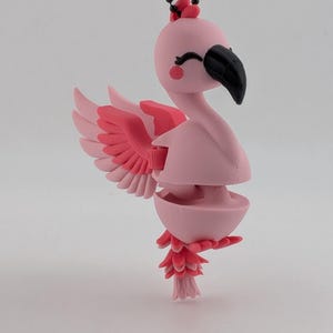 May include: A pink flamingo figurine with a black beak and closed eyes, featuring articulated wings and a segmented body. The flamingo is suspended from a black beaded chain, showcasing a light pink head, darker pink wing accents, and a tail.