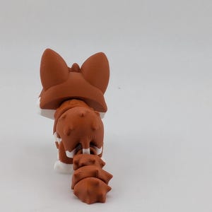 Peut inclure: Une petite figurine de renard articul&eacute;e avec une finition mate dans des tons brun-rouge et blanc. Le renard est repr&eacute;sent&eacute; de dos, mettant en valeur ses oreilles pointues, son dos textur&eacute; et sa queue segment&eacute;e et flexible.