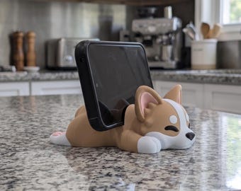 May include: A tan and white corgi-shaped phone stand holding a black smartphone. The dog figure is in a lying position with its head resting on its front paws, featuring pink inner ears, a black nose, and closed eyes. The stand is made of smooth material.