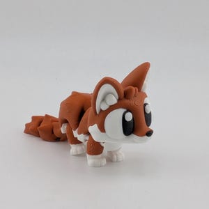 Peut inclure: Une figurine de renard articul&eacute;e imprim&eacute;e en 3D avec un coloris rouille et blanc. Le renard pr&eacute;sente de grands contours d'yeux blancs stylis&eacute;s avec des pupilles noires, des oreilles pointues et une queue segment&eacute;e con&ccedil;ue pour le mouvement.