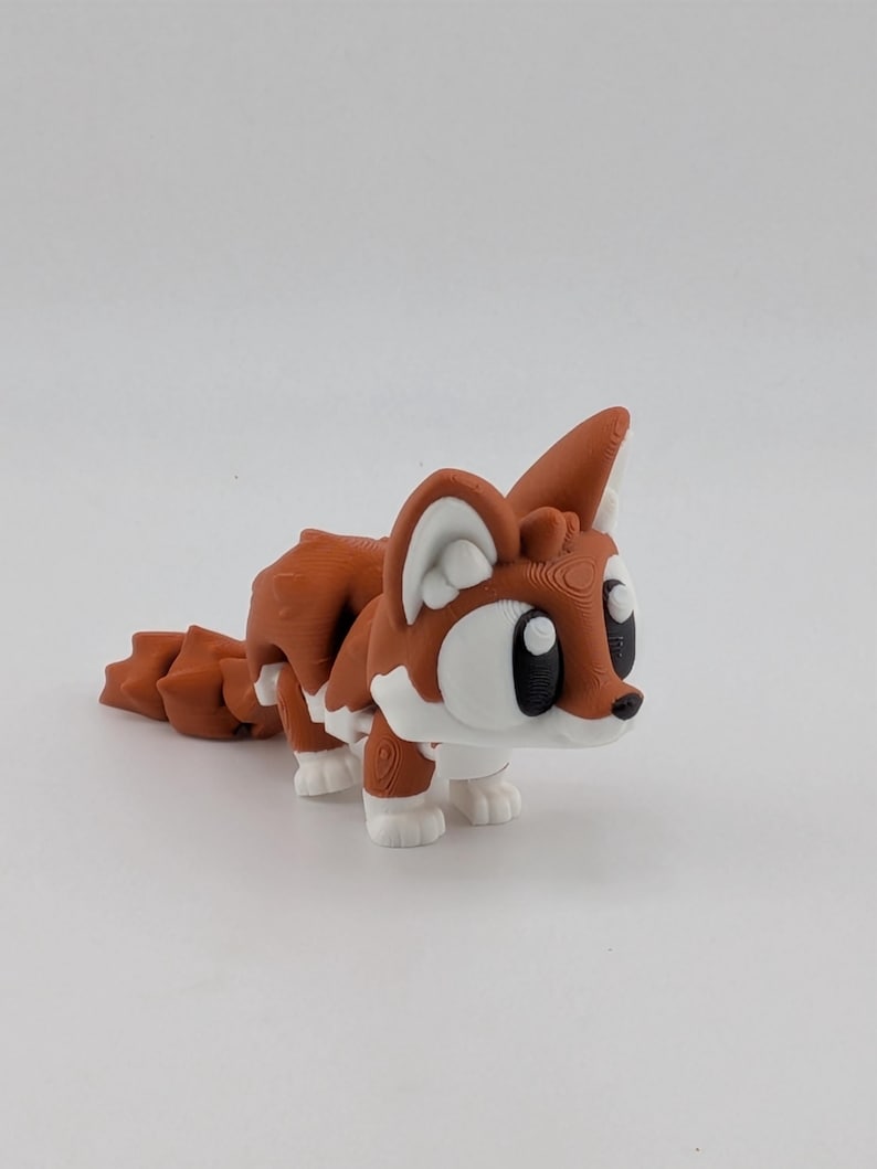 Peut inclure: Une figurine de renard articul&eacute;e imprim&eacute;e en 3D avec un coloris rouille et blanc. Le renard pr&eacute;sente de grands contours d'yeux blancs stylis&eacute;s avec des pupilles noires, des oreilles pointues et une queue segment&eacute;e con&ccedil;ue pour le mouvement.