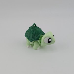 May include: A small, light green turtle figurine with a dark green shell. The turtle has white feet and large eyes with black pupils. The figurine has a loop at the top for hanging.