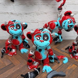 May include: A collection of articulated monkey figurines featuring a vibrant red and light blue color scheme. Each monkey has stitched detailing, silver bolt-like accents on the head, and one eye replaced by a white cross. Several black banana accessories are scattered around the figures.