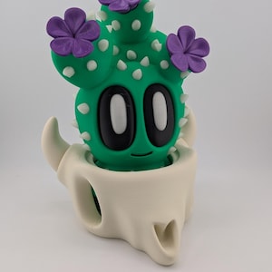 May include: A whimsical green cactus figurine with white spikes and purple flower accents. The cactus has large black eyes and a smiling face. It is set inside a cream-colored skull-shaped holder, creating a unique decorative piece.