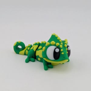 May include: A whimsical, handcrafted chameleon figurine. The reptile is primarily green with yellow spots and accents. It has large, expressive eyes and a curled tail. The figurine is likely made of polymer clay or a similar material.