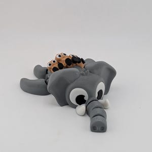 May include: A gray 3D-printed elephant figurine with large white-and-black eyes, white tusks, and a trunk. The elephant has a brown and black object on its back. The figurine is lying on its side.