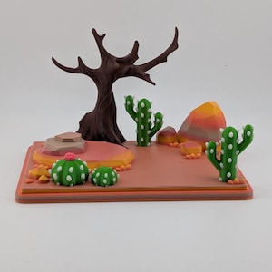 Peut inclure: Un diorama de d&eacute;sert imprim&eacute; en 3D comprenant un arbre brun fonc&eacute; sans feuilles, deux cactus saguaro verts, deux petits cactus ronds verts avec des fleurs roses et plusieurs rochers g&eacute;om&eacute;triques multicolores sur une base rectangulaire.