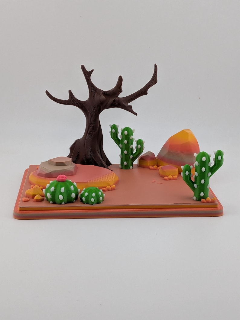 Peut inclure: Un diorama de d&eacute;sert imprim&eacute; en 3D comprenant un arbre brun fonc&eacute; sans feuilles, deux cactus saguaro verts, deux petits cactus ronds verts avec des fleurs roses et plusieurs rochers g&eacute;om&eacute;triques multicolores sur une base rectangulaire.