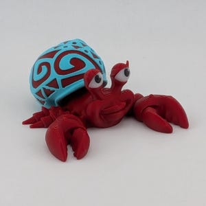 May include: A 3D printed crab figurine with a deep red body and two large eyes on stalks. The crab carries a shell on its back that is dark red with a light blue, raised tribal-style pattern. The figure has two large front claws and multiple legs.