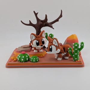 Peut inclure: Un diorama de d&eacute;sert imprim&eacute; en 3D repr&eacute;sentant deux renards stylis&eacute;s marron et blanc avec de grands yeux. La sc&egrave;ne comprend un arbre brun fonc&eacute; sans feuilles, des cactus verts &agrave; points blancs, une petite fleur rose et des rochers orange.