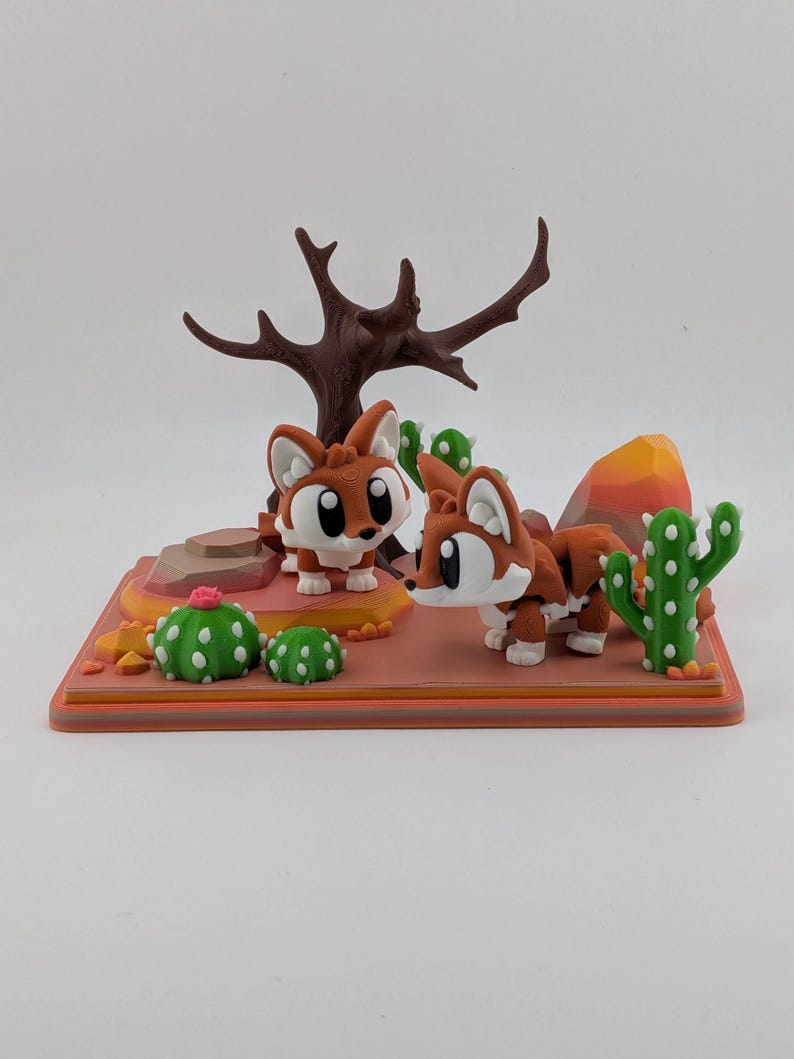 Peut inclure: Un diorama de d&eacute;sert imprim&eacute; en 3D repr&eacute;sentant deux renards stylis&eacute;s marron et blanc avec de grands yeux. La sc&egrave;ne comprend un arbre brun fonc&eacute; sans feuilles, des cactus verts &agrave; points blancs, une petite fleur rose et des rochers orange.