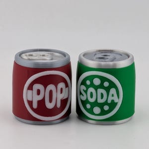 May include: Two cylindrical containers shaped like beverage cans, one red and one green. The red can features a white circular emblem with the text POP, while the green can has a white circular emblem with the text SODA. Both have silver tops.