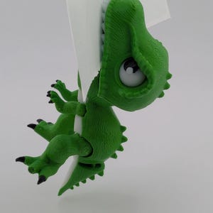 May include: A green 3D-printed dinosaur figurine with a large head, white eyes with black pupils, and sharp white teeth. The dinosaur is depicted in a hanging position, gripping a thin white vertical surface with its front and back claws.