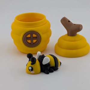 May include: A yellow and black bee figurine with a cartoonish design, next to a yellow beehive-shaped container and a lid. The container has a brown cross-shaped design on the front. The lid has a brown handle. The bee has white wings.