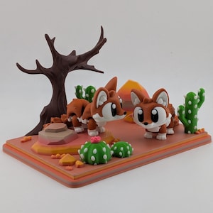 Peut inclure: Un diorama de d&eacute;sert imprim&eacute; en 3D repr&eacute;sentant deux petites figurines de renards marron et blanc. La sc&egrave;ne comprend un arbre brun fonc&eacute; sans feuilles, des cactus verts &agrave; &eacute;pines blanches, une petite fleur rose et des rochers orange.