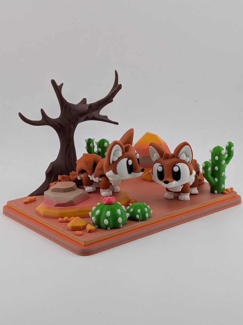 Peut inclure: Un diorama de d&eacute;sert imprim&eacute; en 3D repr&eacute;sentant deux petites figurines de renards marron et blanc. La sc&egrave;ne comprend un arbre brun fonc&eacute; sans feuilles, des cactus verts &agrave; &eacute;pines blanches, une petite fleur rose et des rochers orange.