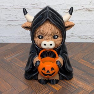 May include: A figurine of a brown Highland cow wearing a black hooded cloak with white horns. The cow has large black eyes and is holding a bright orange plastic pumpkin bucket with a carved jack-o'-lantern face in its front hooves.