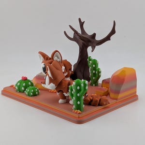Peut inclure: Un diorama imprim&eacute; en 3D repr&eacute;sentant une figurine de renard stylis&eacute;e marron et blanche sur un socle sur le th&egrave;me du d&eacute;sert. La sc&egrave;ne comprend un arbre brun fonc&eacute; sans feuilles, des cactus verts &agrave; points blancs et un rocher ray&eacute;.