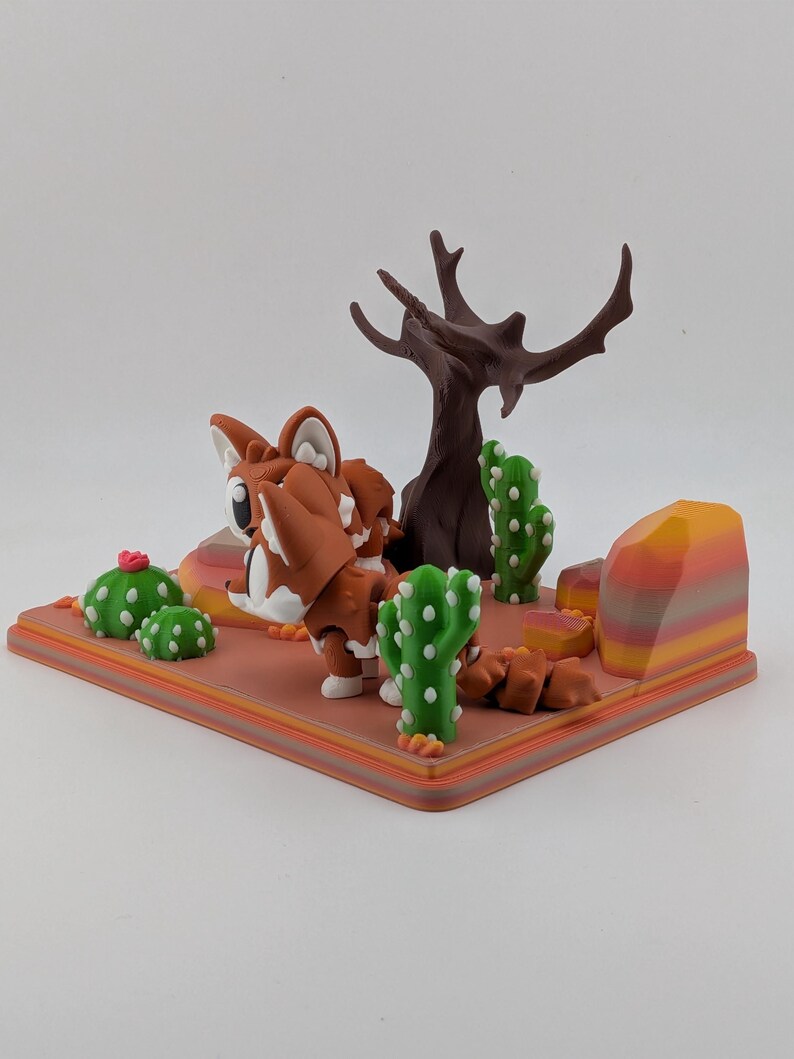 Peut inclure: Un diorama imprim&eacute; en 3D repr&eacute;sentant une figurine de renard stylis&eacute;e marron et blanche sur un socle sur le th&egrave;me du d&eacute;sert. La sc&egrave;ne comprend un arbre brun fonc&eacute; sans feuilles, des cactus verts &agrave; points blancs et un rocher ray&eacute;.