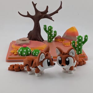 Peut inclure: Deux figurines de renard articul&eacute;es aux corps marron et blanc avec de grands yeux, plac&eacute;es devant un socle de pr&eacute;sentation sur le th&egrave;me du d&eacute;sert avec un arbre marron, des cactus verts &agrave; points blancs et des rochers orange et roses.
