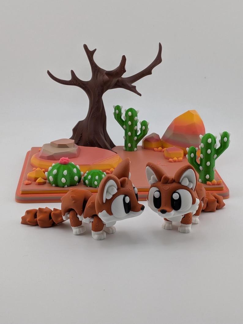 Peut inclure: Deux figurines de renard articul&eacute;es aux corps marron et blanc avec de grands yeux, plac&eacute;es devant un socle de pr&eacute;sentation sur le th&egrave;me du d&eacute;sert avec un arbre marron, des cactus verts &agrave; points blancs et des rochers orange et roses.