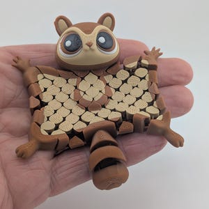 May include: A brown and tan 3D-printed sugar glider figurine with large eyes and outstretched limbs. The glider's body is composed of a series of interlocking shapes, and it is held in a person's hand.
