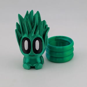 May include: A green 3D-printed figurine of a cartoon-like character with large black eyes and spiky hair, next to a matching green ring. The figurine has a small body and is sitting. The ring is made of three stacked bands.