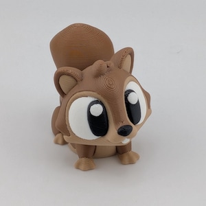 May include: A 3D-printed figurine of a cartoon squirrel. The squirrel has large white eyes with black pupils, a black nose, and a light brown body with darker brown accents. The tail is curled upwards.