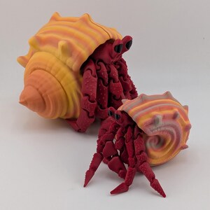 May include: Two articulated hermit crab figures with deep red bodies and textured limbs. Each crab features a spiraled shell in a gradient of yellow, orange, and pink hues. The larger crab is positioned behind the smaller one, both with black eyes.