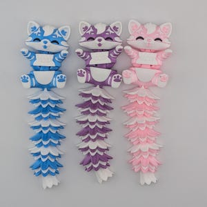 May include: Three colorful articulated fox toys. The toys are blue, purple, and pink, with white accents. Each fox has a segmented tail and is posed with its arms up. The toys are on a plain white background.