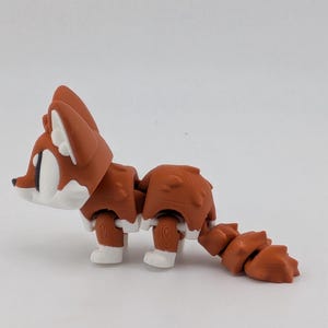 Peut inclure: Une petite figurine de renard articul&eacute;e pr&eacute;sentant un coloris rouille et blanc. La figurine poss&egrave;de une t&ecirc;te stylis&eacute;e avec de grands yeux, un corps segment&eacute; et une queue multi-articul&eacute;e. La texture de surface montre des lignes d'impression 3D.