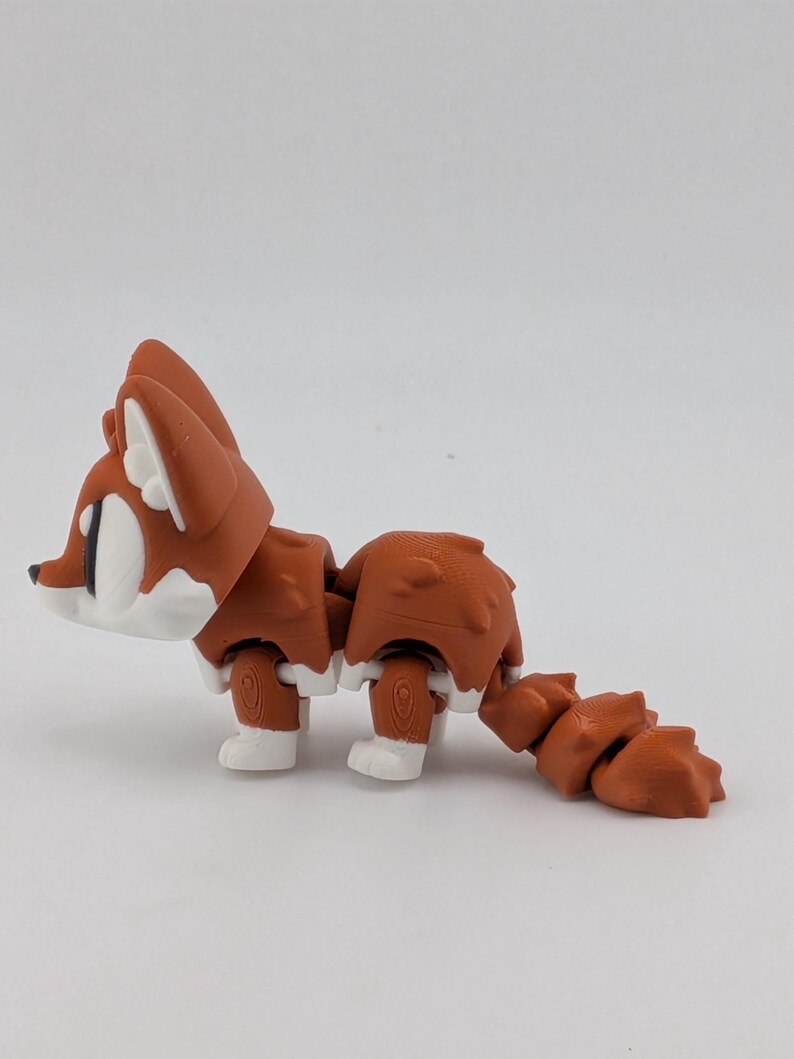 Peut inclure: Une petite figurine de renard articul&eacute;e pr&eacute;sentant un coloris rouille et blanc. La figurine poss&egrave;de une t&ecirc;te stylis&eacute;e avec de grands yeux, un corps segment&eacute; et une queue multi-articul&eacute;e. La texture de surface montre des lignes d'impression 3D.