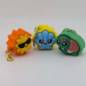 May include: Three small silicone keychain cases featuring cartoon characters. One is an orange sun with black sunglasses, one is a yellow and blue seashell with a smiling face, and one is a green turtle with a pink shell. Each has a ball chain.