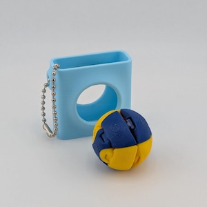 May include: A light blue protective case with a silver chain next to a yellow and blue puzzle ball. The case has a circular opening. The puzzle ball is spherical and has a segmented design.
