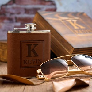以下が含まれることがあります： イニシャル「K」と「KEVIN」が刻まれた茶色の革製フラスコ。茶色のレンズのゴールドフレームサングラスがフラスコの隣にあります。「K」の文字が入った木箱が背景にあります。