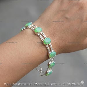 May include: A silver bracelet featuring oval, light green gemstones. The bracelet is composed of silver links connecting the gemstones. The gemstones are set in a silver bezel setting. The bracelet is on a wrist.