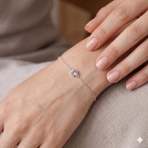 May include: A delicate silver chain bracelet featuring a single, round, light purple gemstone set in a simple metal bezel. The bracelet is secured with a thin, polished silver link chain that wraps around the wrist, highlighting the gemstone.