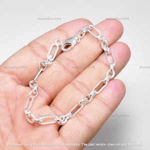 Puede incluir: Una pulsera de plata con un diseño de cadena de eslabones rectangulares. La pulsera está hecha de metal brillante y presenta un cierre de mosquetón. La pulsera se muestra sobre un fondo claro.