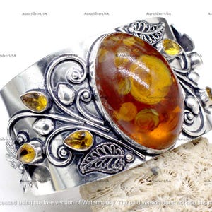 May include: A wide silver cuff bracelet featuring an oval amber stone as the centerpiece. The bracelet is adorned with intricate leaf and floral designs, and smaller amber-colored stones. The bracelet is displayed on a textured surface.