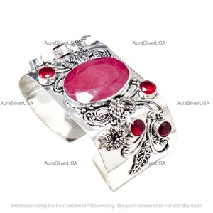 May include: A wide silver cuff bracelet with an oval, large, ruby-colored gemstone in the center, surrounded by smaller ruby-colored stones. The bracelet has an ornate, floral design and the text "AuraSilverUSA".