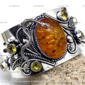 Op de afbeelding: Een brede zilveren manchetarmband met een ovale barnsteen in het midden, geaccentueerd door een gedetailleerde zilveren slang en geelgroene edelstenen. De armband heeft een sierlijk ontwerp met bloemen- en krulwerkdetails. De tekst "AuraSilverUSA" is zichtbaar.