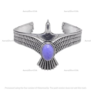 Pode incluir: Uma pulseira de punho prateada com um design de águia. A águia tem as asas abertas e uma pedra ovalada roxa clara. A pulseira tem detalhes de penas e o texto "AuraSilverUSA".