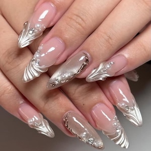 May include: A set of long, almond-shaped artificial nails. The nails feature a nude pink base with clear tips. Some nails have silver, fan-shaped accents, pearls, and silver rhinestones. Other nails have a silver glitter design and gold trim.