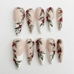 May include: A set of ten almond-shaped press-on nails featuring a nude and white base with hand-painted green holly leaves, dark green foliage, and small red gemstone accents. Each nail is decorated with white polka dots and botanical motifs.
