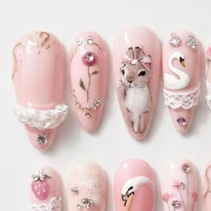 May include: A set of light pink press-on nails featuring intricate designs including a white lace trim, a painted rose with a central crystal, a detailed rabbit illustration with a bow, a white swan figure, a strawberry charm, and small rhinestones.